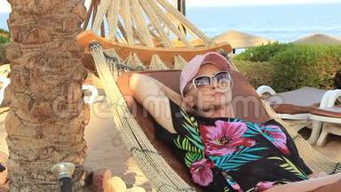 美丽的年轻女子戴着帽子，在阳光明媚的<strong>夏日</strong>棕榈树的树荫下，躺在<strong>海滨</strong>的吊床上