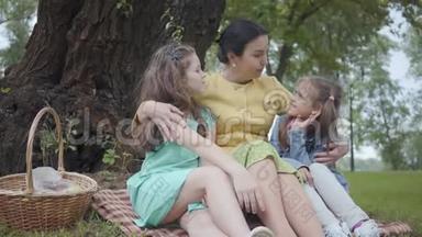 肖像优雅的高级妇女坐在公园树下的草地上，拥抱着两个可爱的孙女，讲述着