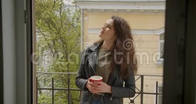 穿着皮夹克的漂亮黑发女孩站在阳台上，欣赏着绿色的城市景色，喝着热茶，充满了梦幻和梦幻