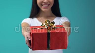 开朗的女孩展示礼品盒到相机，隔离在蓝色背景，假日