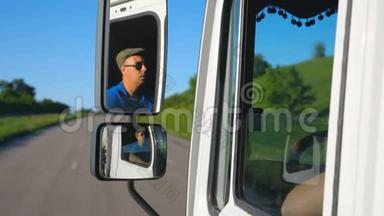卡车司机戴着太阳镜，在移动汽车的侧面镜子里反射。 男子驾驶卡车通过