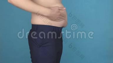 一个年轻人在蓝色背景下拍着肥胖的胖肚子，肥胖和肥胖问题的概念