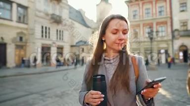 女人手里拿着一个<strong>保温杯</strong>，在夕阳下用智能手机沿着一条老街走着。 通信、社交网络