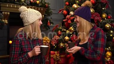 两个穿着红色格子衬衫的女孩<strong>坐在</strong>圣诞<strong>树下</strong>，戴着温暖的白色和紫色的帽子，谈论她们的事情。