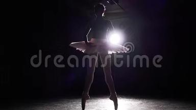 年轻的苗条芭蕾舞演员练习芭蕾。 在演播室的黑色背景上，芭蕾舞演员的剪影被孤立在聚光灯下
