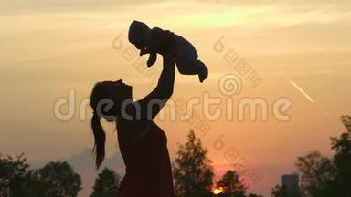 日落剪影：年轻的母亲抱着她的孩子在城市公园站在夕阳面前，生动。