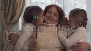 幸福的奶奶坐在靠窗的椅子上，两个女孩站在她身边.. 拥抱美丽的孙女