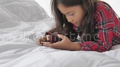 可爱的女孩躺在床上，看着智能手机和社交媒体娱乐。 人们的生活方式和教育。 儿童和儿童