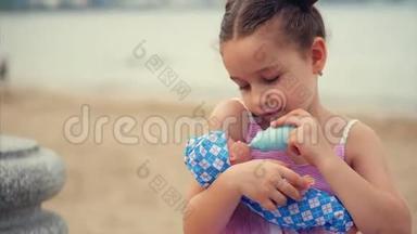 可爱的小女孩用玩具瓶子喂她最喜欢的娃娃。 幼儿在海边的公园里玩娃娃.. 开心快乐