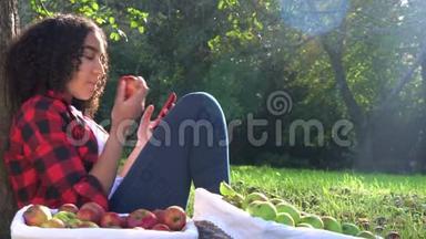一个不分种族的少女，年轻女子坐在树旁，用她的手机智能手机吃苹果