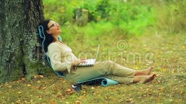 一个戴眼镜的女人赤脚<strong>坐在</strong>公园的<strong>树下</strong>，用笔记本电脑工作。