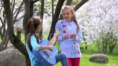 快乐和积极的两个学校女朋友在自然公园玩音乐和唱歌。 一个年轻女孩弹吉他，另一个女孩拍手