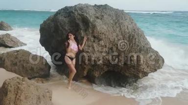 慢动作，漂亮的年轻女孩，穿着泳衣，站在大海和海浪背景下的大石头附近，稳重的。