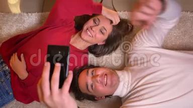 图为一对年轻的白种夫妇头对头趴在地板上，用智能手机拍自拍