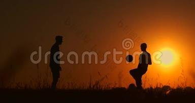 两个男孩在日落时踢足球。 日落时孩子们玩球的剪影。 幸福家庭的概念