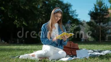 学习笔记的女孩。 年轻女子坐在公园的草地上，拿着一本打开的笔记本，远远地看着，