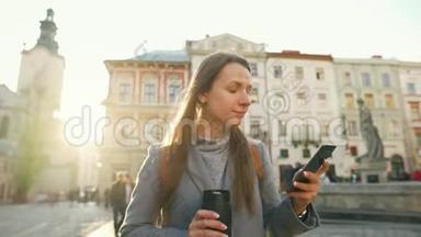 女人手里拿着一个保温杯，在夕阳下用智能手机沿着一条老街走着。 通信、社交网络