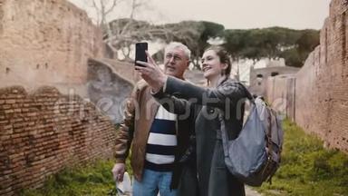 兴奋快乐的高级男人和微笑的欧洲年轻女子在意大利奥斯蒂亚的旧废墟附近自拍度假之旅。