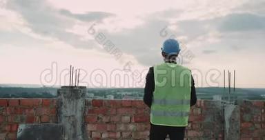 年轻的工程师带着安全帽和高能见度背心，从后面拍屋顶建筑背景的照片