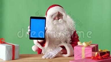 笑容可掬的圣诞老人展示平板电脑，相机里有蓝色屏幕，桌子上有礼物，背景是彩色的。