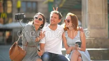 三个快乐的朋友在使用智能手机拍摄自拍户外时，一边拍照一边拍照。 两个漂亮的女孩和一个