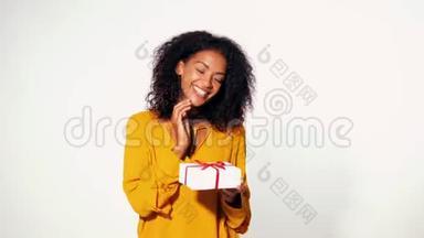 可爱的非裔美国年轻女子，穿着黄色的秋装上衣，手里拿着带有红色丝带的礼品盒，在白色的墙壁背景上鞠躬