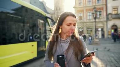 年轻的女人手里拿着一个<strong>保温杯</strong>，在日落时用智能手机沿着一条老街走着。 交流、社会