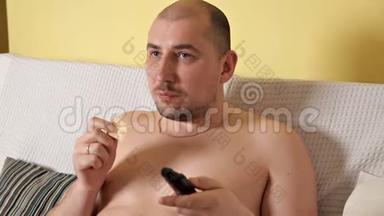 懒洋洋的，秃头的，胖乎乎的，没有外套的胖子正在看电视，在沙发上喝着薯片的可口可乐。 胖子