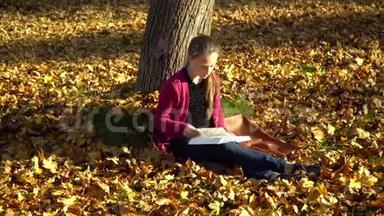 秋天森林里的一棵树<strong>下</strong>坐着一个年轻漂亮的女孩。 一个女孩在读一本书。 她很细心
