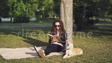 有魅力的女学生正在看书，坐在城市公园树<strong>下</strong>的格子上，小狗躺在附近，享受着快乐