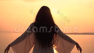 一个穿着海滩礼服的女孩站在夕阳背景上，双手朝不同的方向伸展。 慢动作。 高清高清
