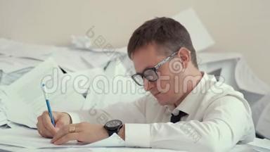 一<strong>个</strong>戴眼镜的疲惫的年轻人坐在一堆文件和签名文件里。 他手臂上有<strong>几个</strong>小时，他累了，