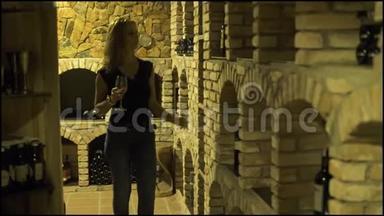 带着红酒酒杯的年轻女人在地窖里看着瓶子。 女子酿酒师在传统酒窖品尝红酒。