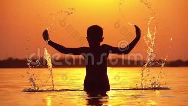日落时分男孩的<strong>剪影</strong>举起双手，创造水的飞溅。 慢动作