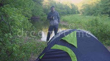 一个年轻人从帐篷和森林河附近的<strong>保温杯</strong>里喝茶
