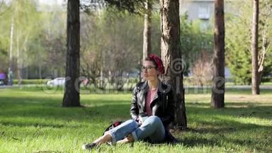 美丽年轻时尚的女人坐在公园的一棵树下，喝着一杯咖啡。 大自然中的外卖咖啡。 时尚时尚时尚