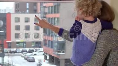 女人透过窗户向孩子展示雪花。 冬天降雪