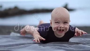 婴儿趴在海边的黑沙上，笑着看着摄像机
