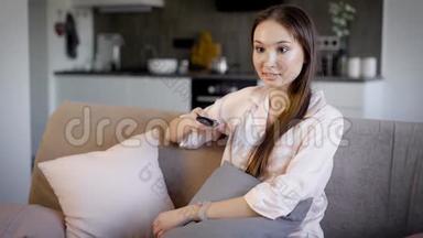 漂亮的黑发女人在度假时躺在客厅的沙发上，通过遥控器切换<strong>电视频道</strong>