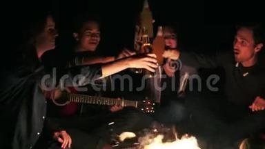 一群年轻人在篝火旁喝着啤酒，深夜喝着欢呼声叮叮作响的瓶子。 慢镜头