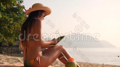 年轻的混合种族旅游女孩使用移动平板小猫和日光浴在热带海滩。 4K，慢动作。 普吉岛