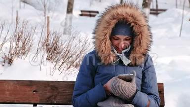 白天雪天，特写女郎坐在长凳上，在城市的冬季公园里浏览手机