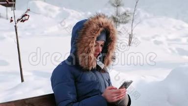 白天雪天，特写女郎坐在长凳上，在城市的冬季公园里浏览手机