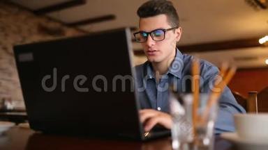 英俊的自由职业者商人戴眼镜，勤奋地在咖啡馆的笔记本电脑上工作。 男子键盘打字和搜索新