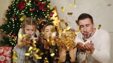 年轻幸福的四口之家坐在圣诞树旁，吹着金色的纸屑。 可爱的母亲，父亲和两个
