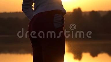 忧郁的女人在夕阳西下的湖岸边弯下腰