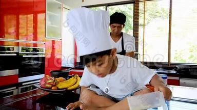 泰国私人厨师烹饪，他的小儿子在厨师`的帽子附近坐在桌子上，在一个现代风格的家庭厨房。 孩子在吃东西，