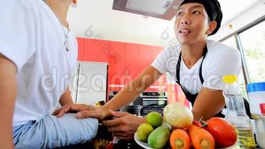 泰国私人厨师烹饪，他的小儿子在厨师`的帽子附近坐在桌子上，在一个现代风格的家庭厨房。 孩子在吃东西，