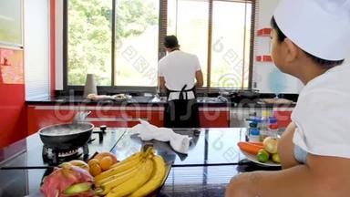 泰国私人厨师做饭，他的小儿子戴着厨师`帽子，坐在桌子上，有一个现代风格的家庭厨房