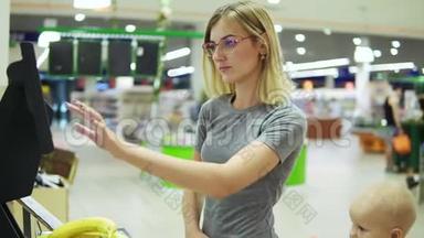年轻的母亲在超市里用自助式电子秤称香蕉，而她可爱的孩子则坐在超市里